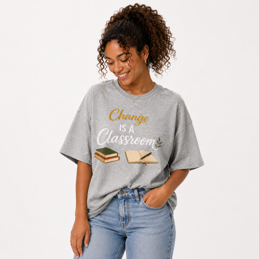 A woman wearing a heather gray graphic t-shirt with the text 'Change IS A Classroom' in gold and white, paired with light blue jeans, against a plain white background.