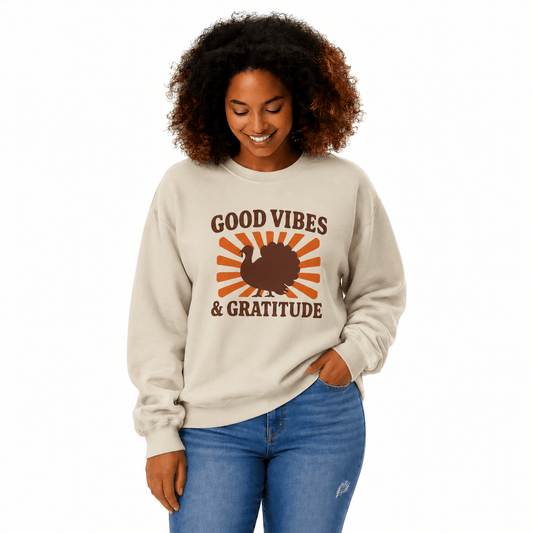 A woman with curly brown hair wearing a beige crewneck sweatshirt with a 'Good Vibes & Gratitude' Thanksgiving graphic, paired with blue jeans, against a plain white background.