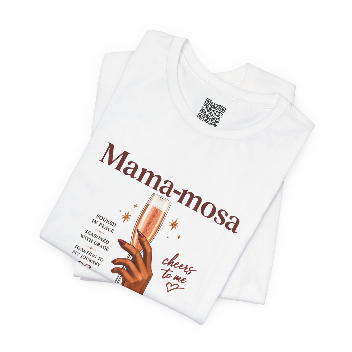 A folded white t-shirt with a graphic print that reads 'Mama-mosa' in dark brown text, featuring an illustration of a hand holding a glass of rosé wine and the phrase 'cheers to me' in cursive. A QR code is printed inside the collar.