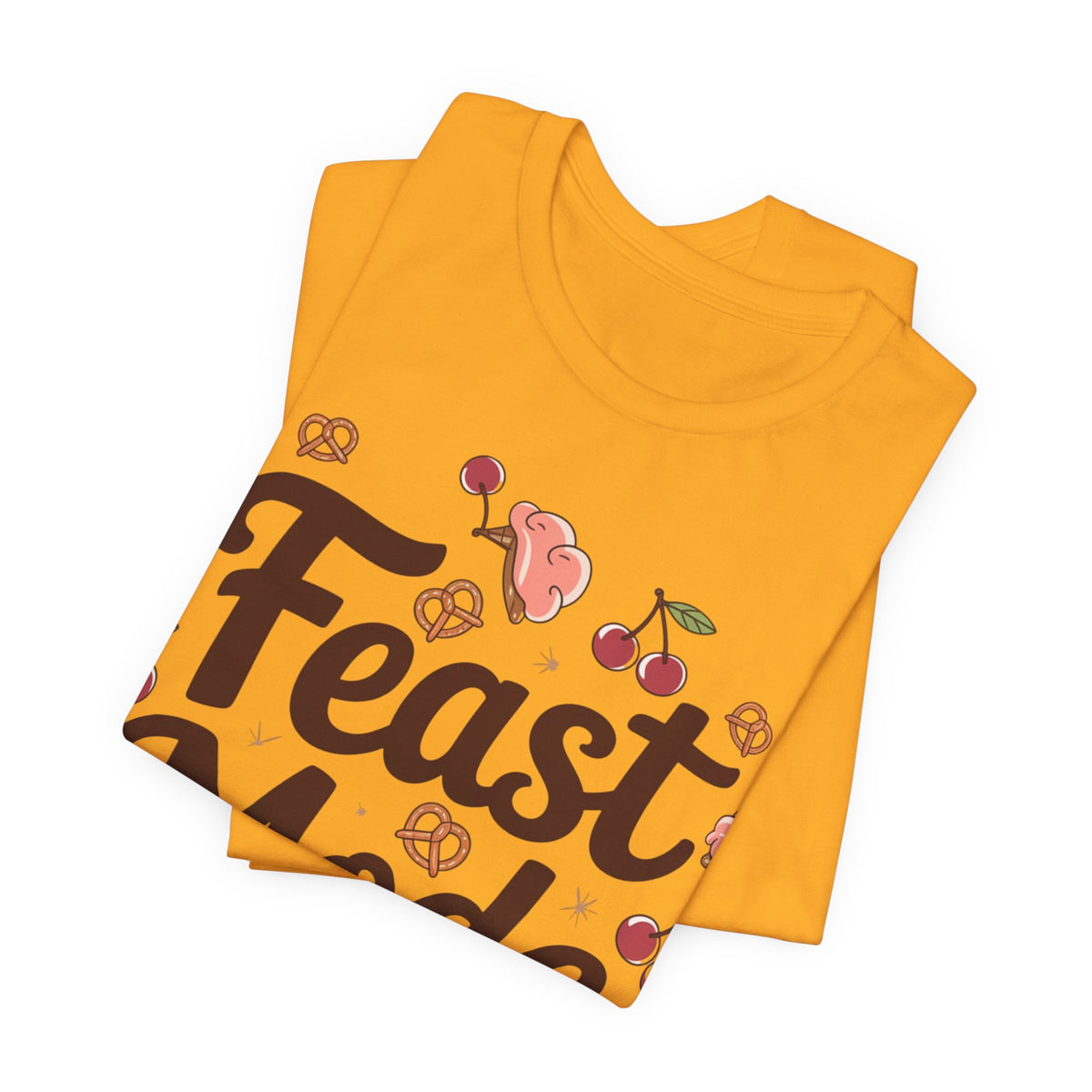 A folded gold t-shirt with a graphic print featuring the word 'Feast' in dark brown script, pretzels, cherries, and a pink donut, displayed on a clean white background.