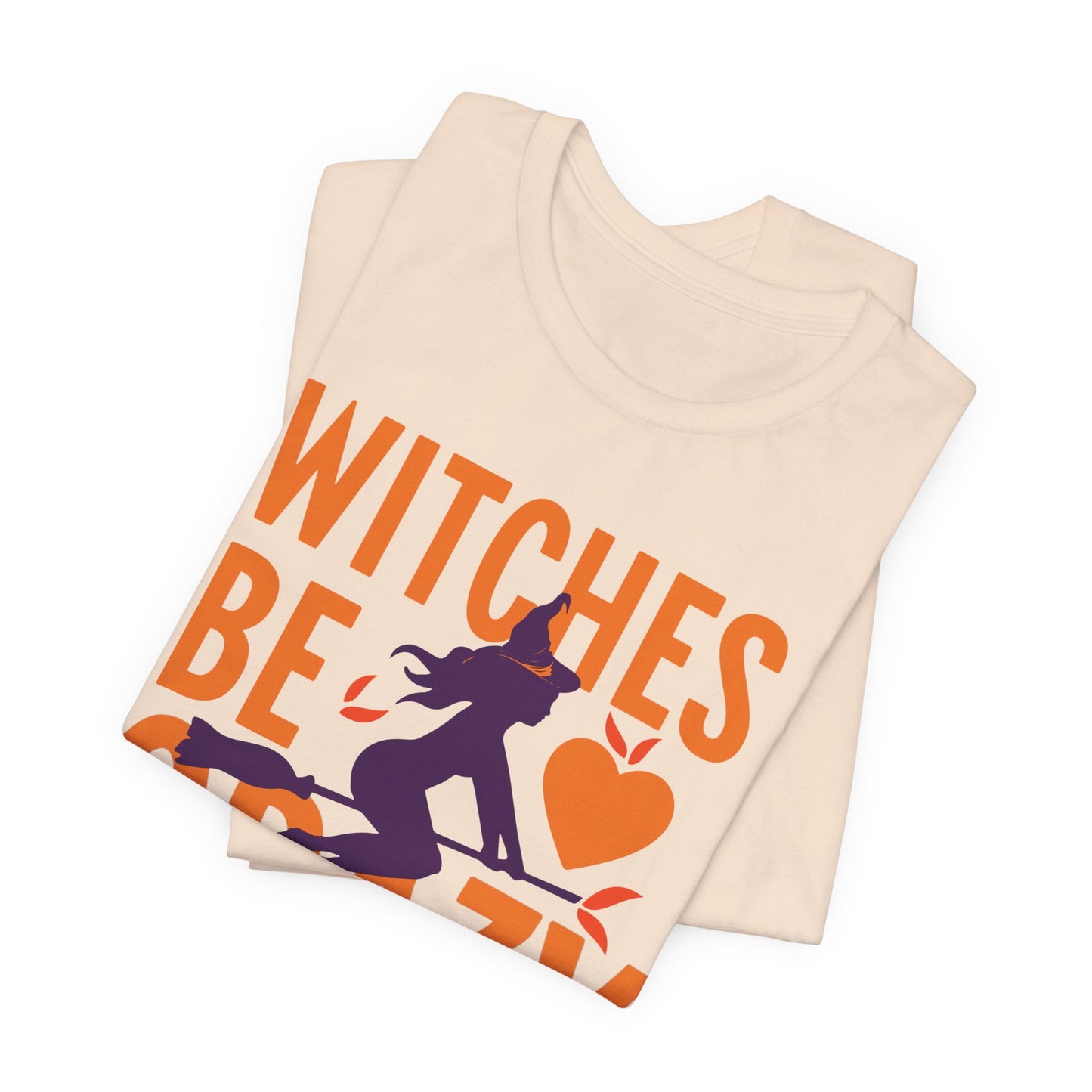 A folded cream-colored t-shirt with a Halloween graphic that reads 'WITCHES BE' in orange, featuring a purple witch riding a broomstick next to an orange heart, shown on a plain white background.