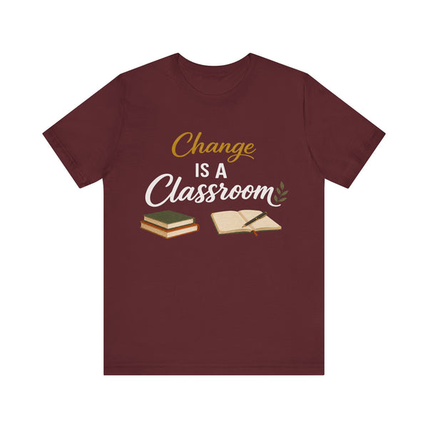 A maroon short-sleeve crewneck t-shirt with a graphic on the chest that reads 'Change IS A Classroom' in white and gold lettering, accompanied by illustrations of stacked books and an open notebook, displayed on a solid white background.