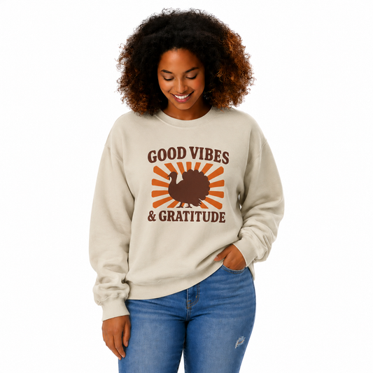 A woman with curly brown hair wearing a beige crewneck sweatshirt with a 'Good Vibes & Gratitude' Thanksgiving graphic, paired with blue jeans, against a plain white background.