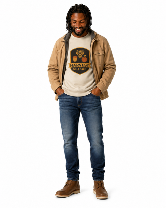 A man modeling a beige jacket over a cream graphic t-shirt, blue jeans, and brown boots against a plain white background.
