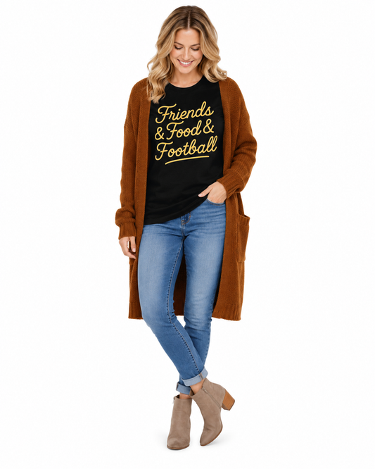 A woman modeling a brown open-front cardigan over a black graphic t-shirt that reads 'Friends & Food & Football' in yellow, paired with blue jeans and taupe ankle boots, on a solid white background.