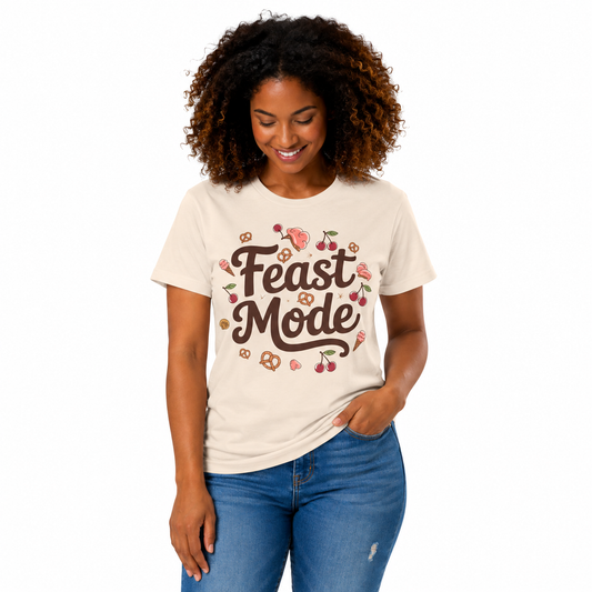 A woman with curly brown hair wearing a cream-colored t-shirt with a 'Feast Mode' graphic and blue jeans, standing against a plain white background.