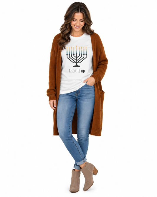 A woman modeling a white graphic t-shirt with a menorah print and the text 'Light it up', styled with a brown open-front cardigan, blue jeans, and taupe ankle boots against a plain white background.