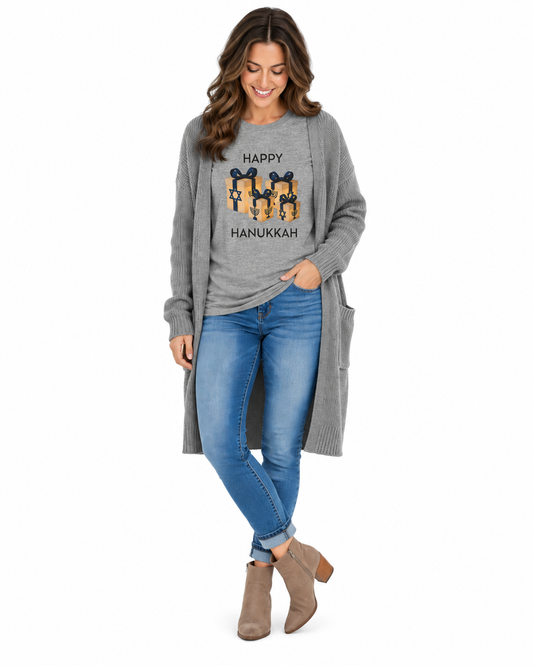 A woman modeling a long-sleeve gray knit cardigan with a 'Happy Hanukkah' graphic featuring cartoon figures, paired with blue jeans and taupe ankle boots, on a solid white background.