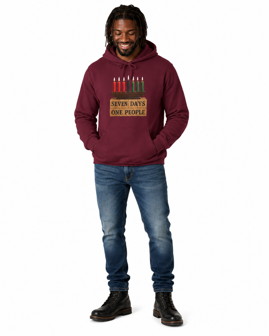 A man wearing a maroon pullover hoodie with a Kwanzaa graphic that reads 'SEVEN DAYS ONE PEOPLE' with seven candles, paired with blue jeans and black boots, standing against a plain white background.