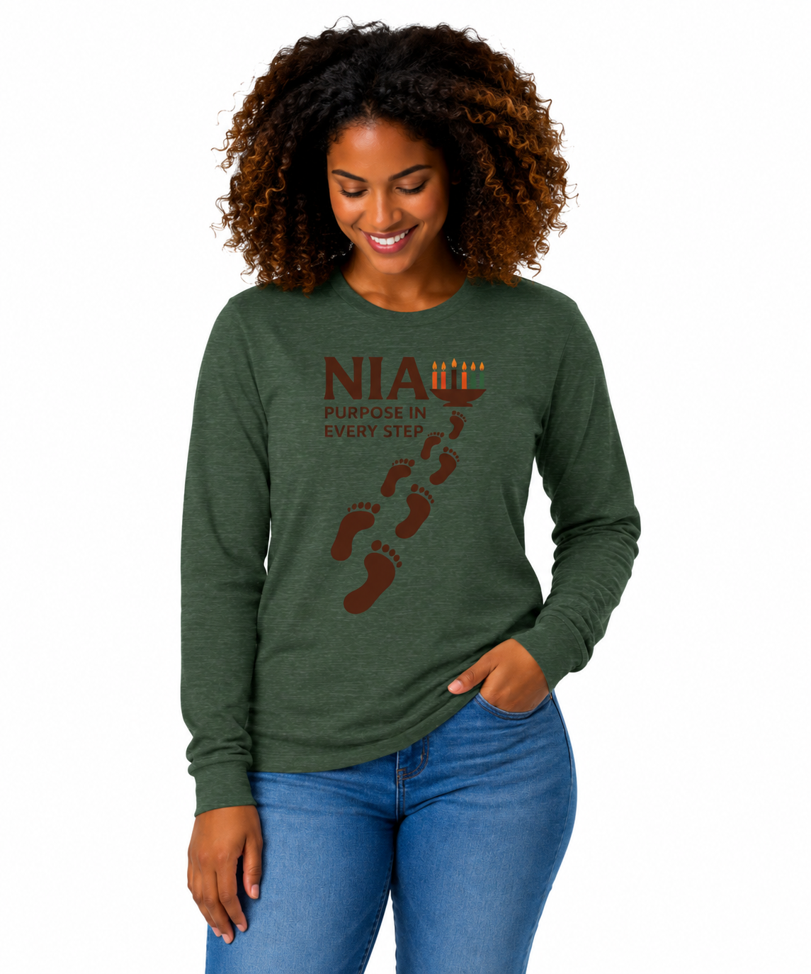 A woman with curly brown hair wearing a heathered forest green long-sleeve t-shirt with a brown and orange graphic that reads 'NIA PURGHELGIN EVERY STEP' with footprints, paired with blue jeans, against a plain white background.