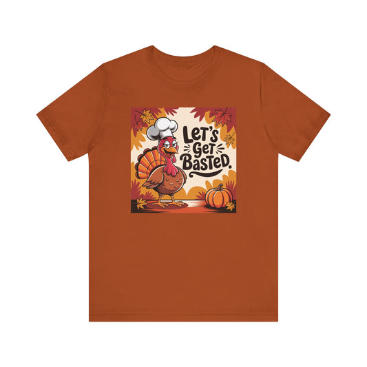 An orange short-sleeve crewneck t-shirt featuring a central graphic of a cartoon turkey wearing a chef's hat with the text 'Let's Get Basted.' in autumn colors, displayed on a solid white background.