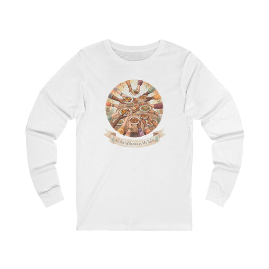 A white long-sleeve crewneck t-shirt featuring a circular graphic on the chest depicting a multi-colored Passover Seder table with hands reaching toward food, and a banner below reading 'All Are Welcome at My Table'.