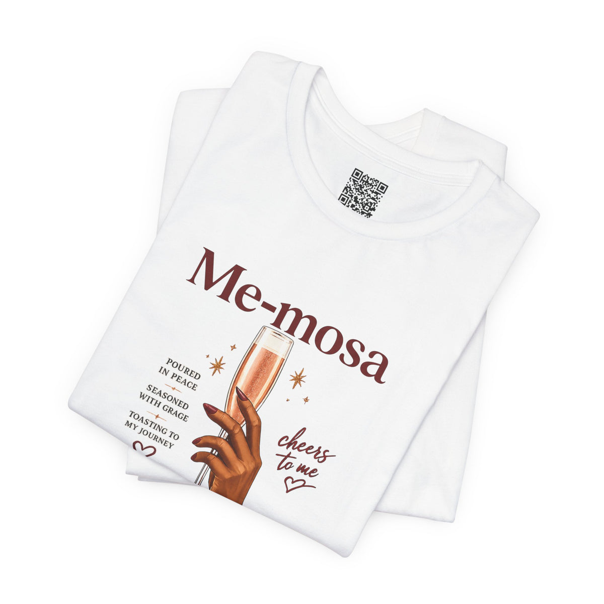 A folded white t-shirt with a graphic print that reads 'Me-mosa' and 'cheers to me' in brown lettering, featuring an illustration of a hand holding a champagne flute. A QR code is visible on the inner neck label.