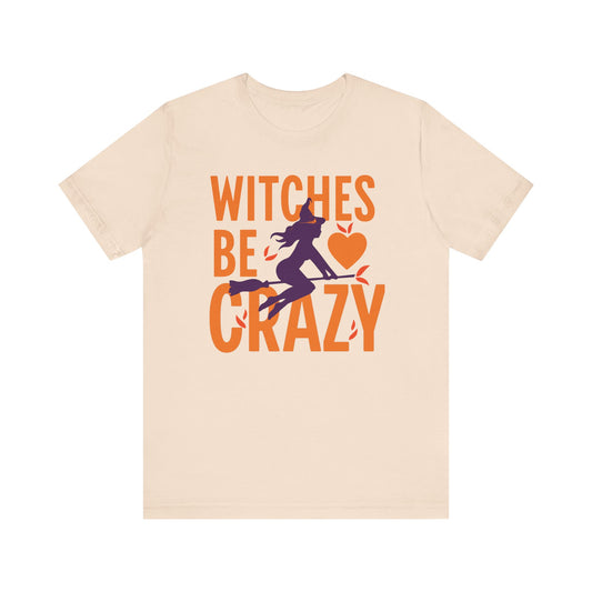 A light beige crewneck t-shirt featuring a Halloween graphic on the front with the text 'WITCHES BE CRAZY' in orange, a purple witch silhouette, and an orange heart.