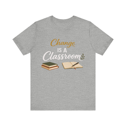 A heather gray short-sleeve t-shirt with a graphic print on the front that reads 'Change IS A Classroom' in white and gold lettering, accompanied by illustrations of stacked books and an open notebook with a pen.