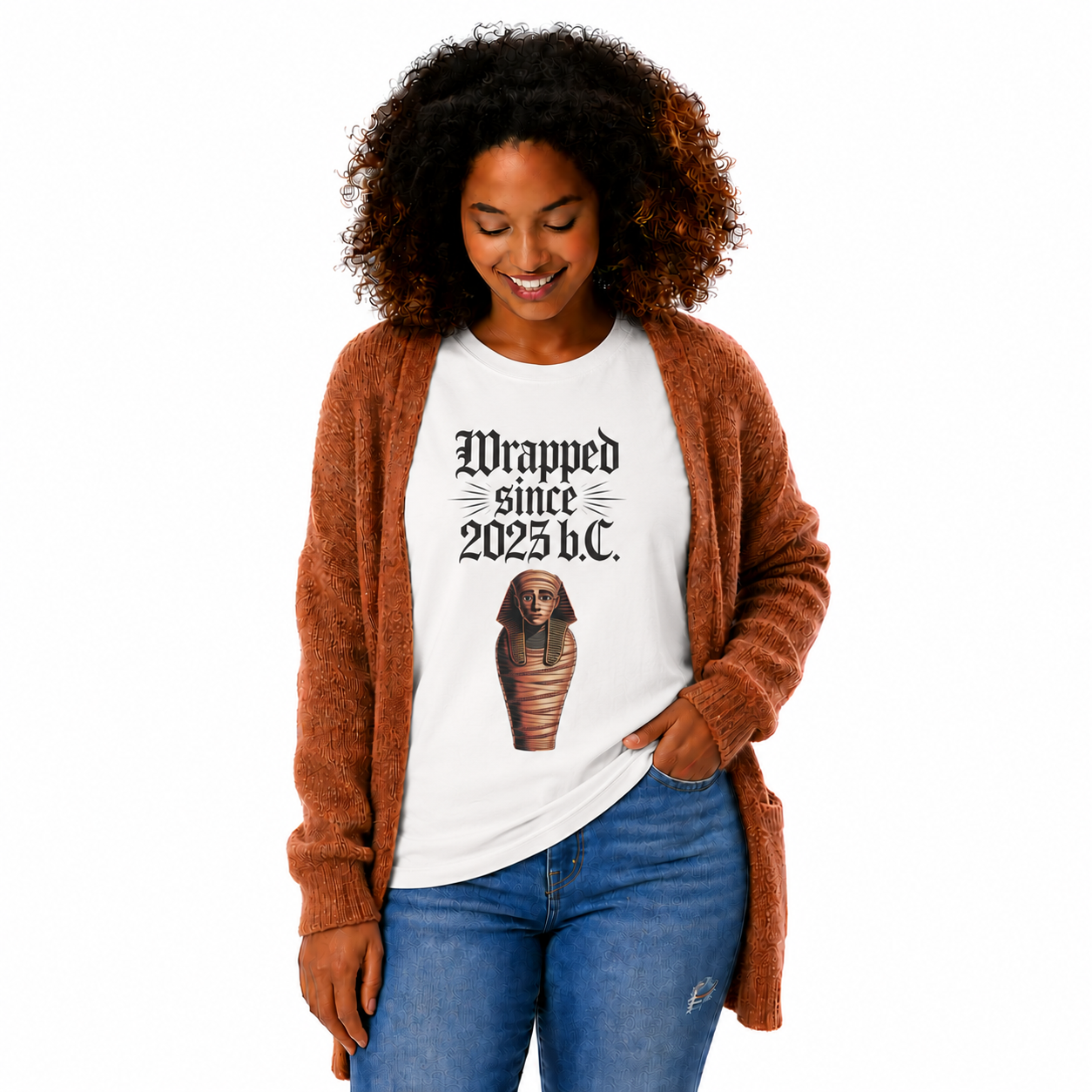 A woman wearing a white graphic t-shirt with a black gothic text print and an Egyptian mummy design, layered under a rust-brown fuzzy cardigan and paired with blue jeans, set against a plain white background.